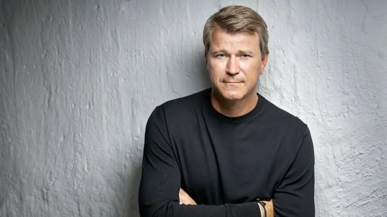Anders Jensen, en man med blont hår och blå ögon, bär en svart tröja och står med armarna i kors mot en vit, strukturerad vägg.