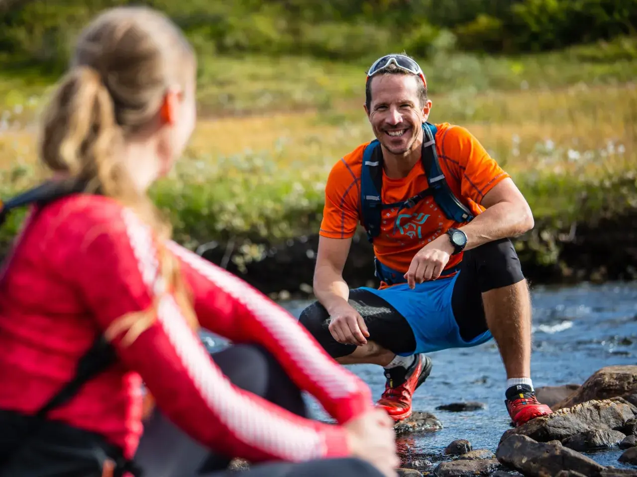 Två löpare i gorp core-stil vid en bäck. Mannen ler glatt i orange tröja och blå shorts.