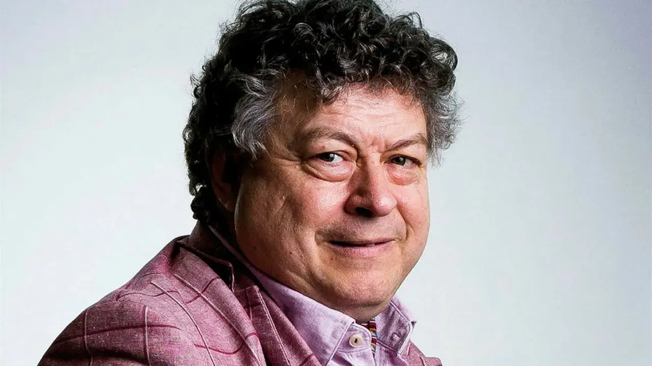 Rory Sutherland, med sitt karaktäristiska lockiga hår och ett vänligt leende, bär en rutig kavaj.