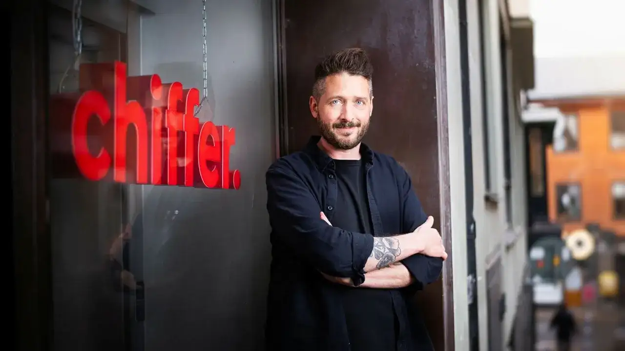 Gary Engman, med tatueringar och skägg, står med armarna i kors utanför en lokal med röd text som lyder "chiffer".