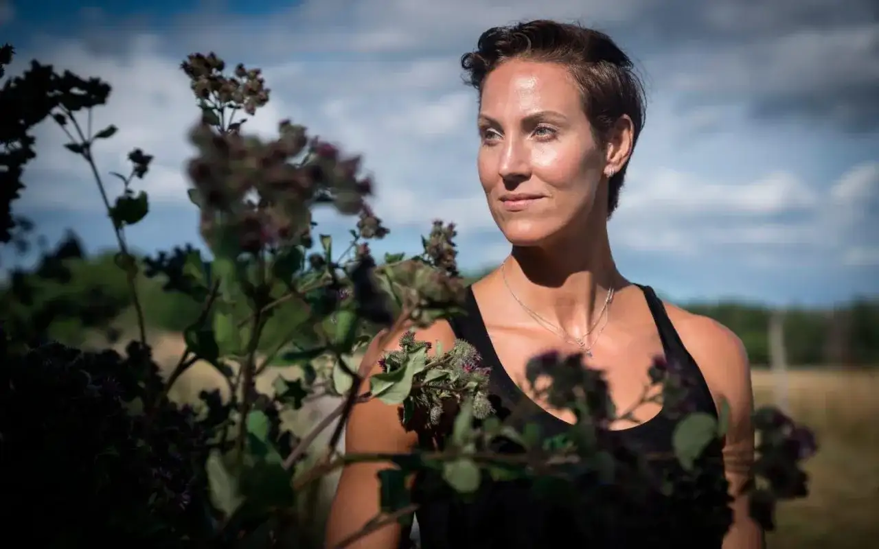Elin Kjos, med kortklippt hår, blickar ut över ett fält. Blommor i förgrunden skapar en naturlig inramning.