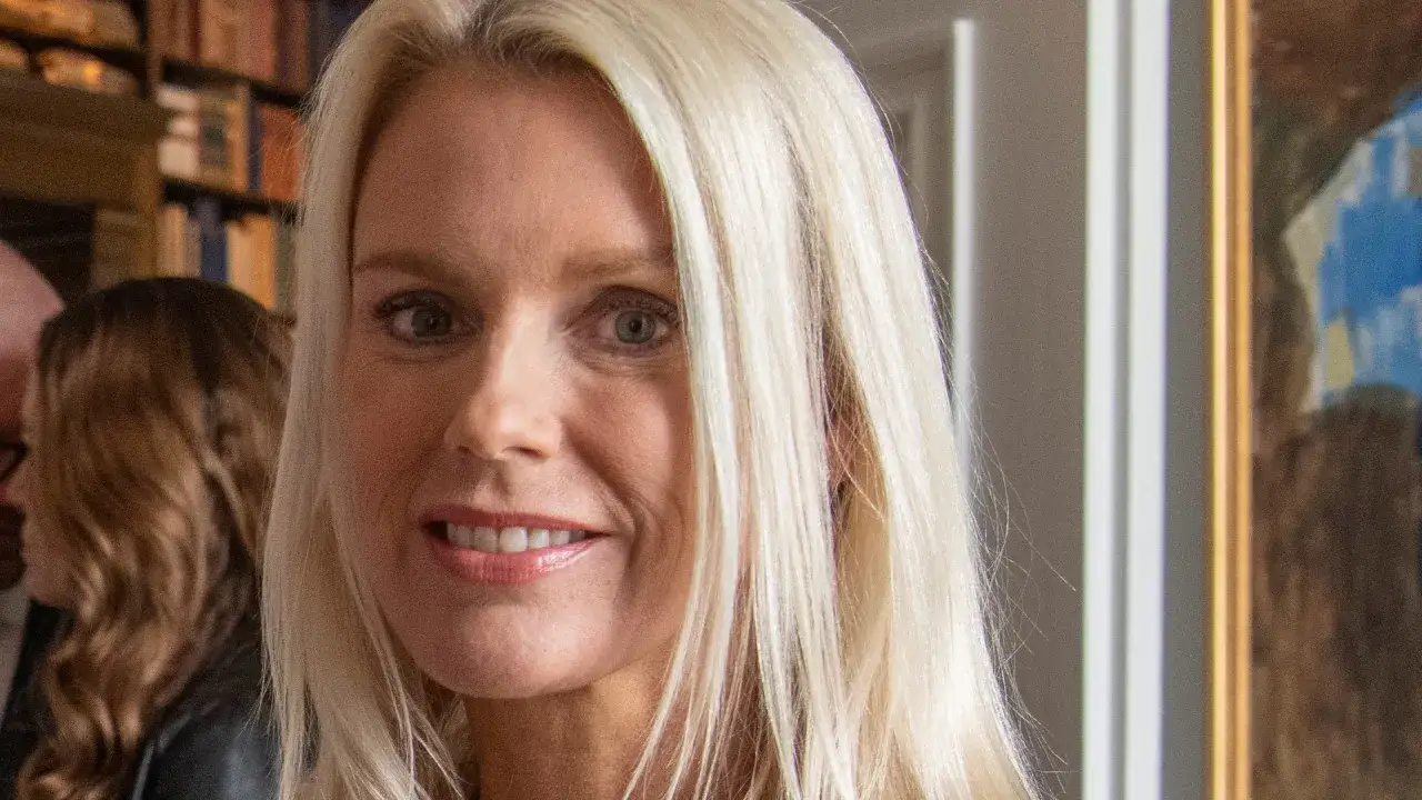 Stina Bergfors ler mot kameran, med sitt blonda hår fallande över axlarna. I bakgrunden syns bokhyllor och en tavla.