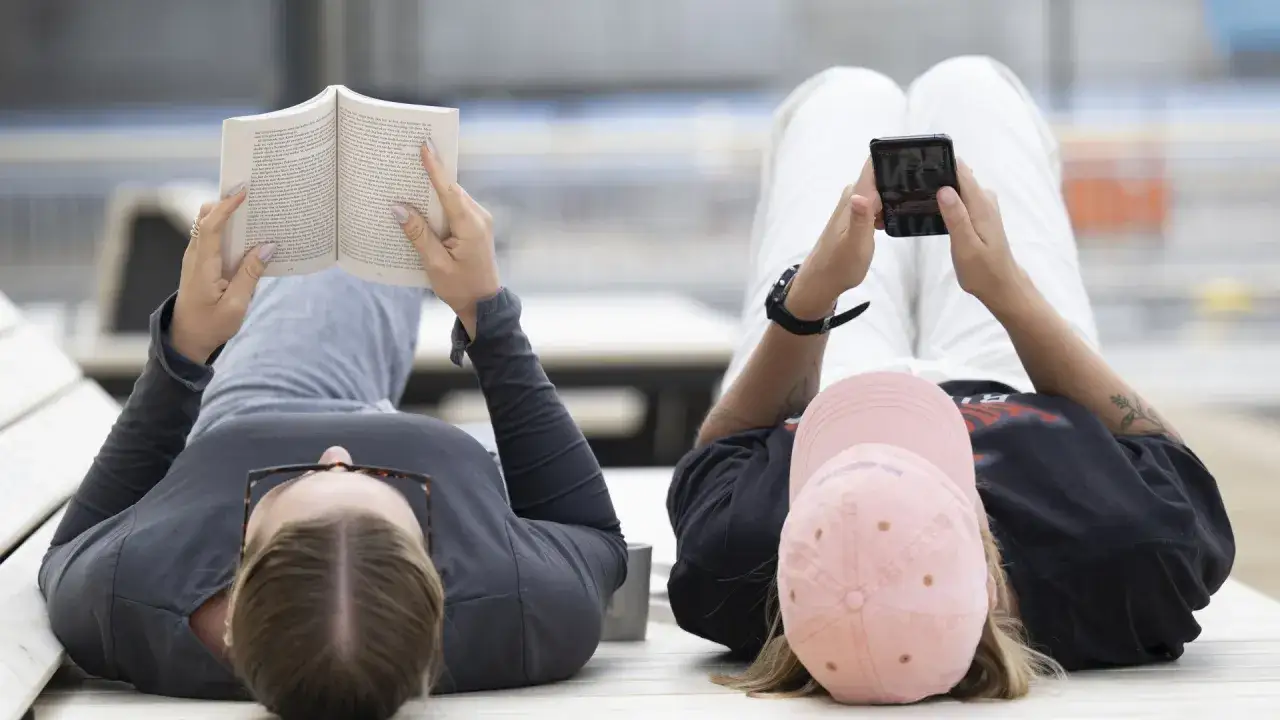 Två personer vilar på en bänk, en läser en bok, den andra tittar på sin mobil. Människor människor människor njuter av lugnet.