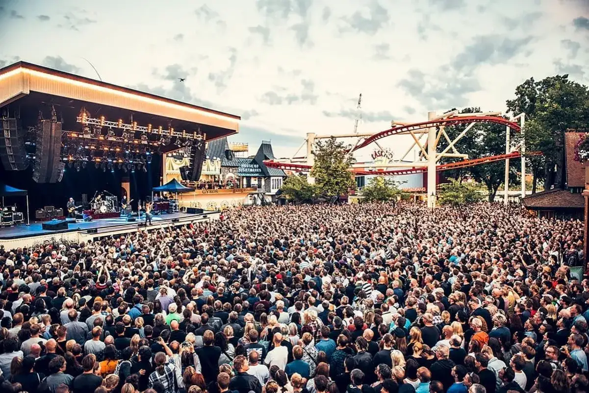 Stor publik p&aring; Gr&ouml;nan. En konsert p&aring; Gr&ouml;na Lund med en enorm folkmassa som njuter av musiken och n&ouml;jesparkens attraktioner i bakgrunden.