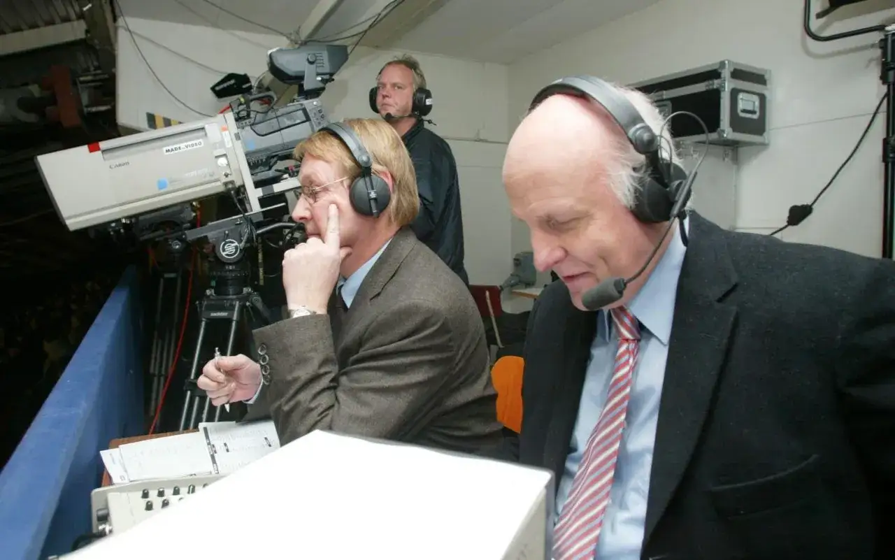 Arne Hegerfors, med headset, sitter vid ett bord med papper. En kameraman st&aring;r bakom.