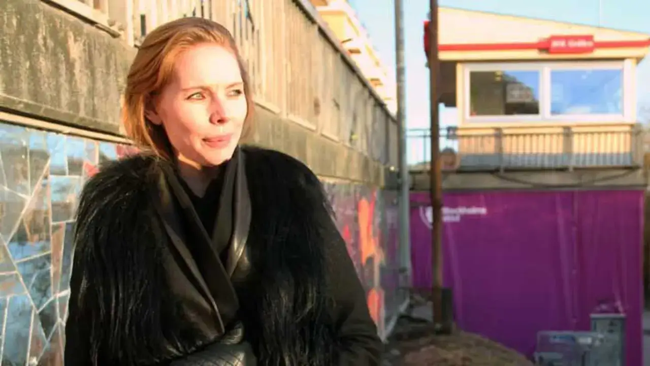 Karin Winther i en svart p&auml;lsjacka framf&ouml;r en v&auml;gg med mosaik och en lila presenning med texten 