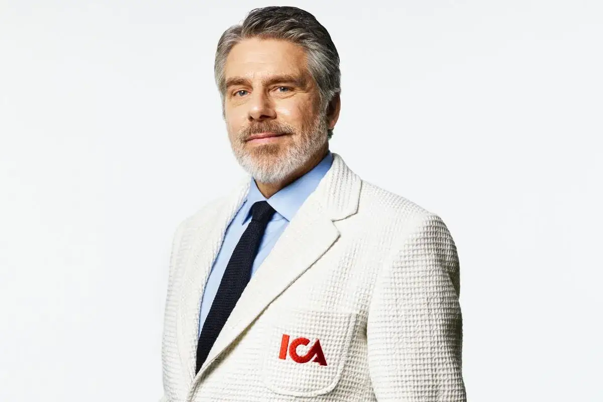 Man i vit kavaj med ICA-logga på bröstet. Han har grått hår och skägg, blå skjorta och mörk slips. En ny ICA-profil.