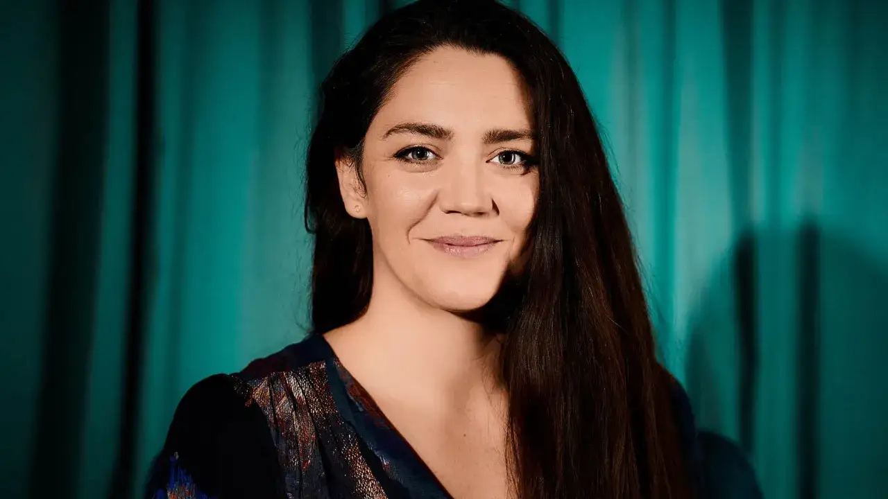 Jenny Nordlander ler mot kameran, med m&ouml;rkt h&aring;r som faller &ouml;ver axeln. Bakgrunden &auml;r en turkos gardin.
