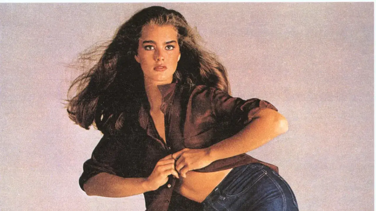 Brooke Shields i ikoniska Calvin Klein-jeans. Hennes l&aring;nga, m&ouml;rka h&aring;r fl&ouml;dar medan hon kn&auml;pper upp sin bruna skjorta.