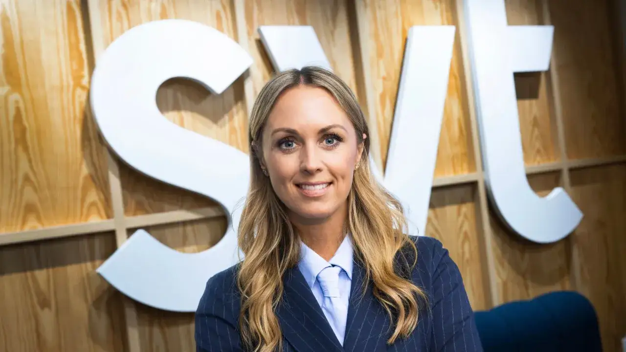 En kvinnlig programledare för SVT ler mot kameran, klädd i kavaj och ljusblå skjorta med slips.