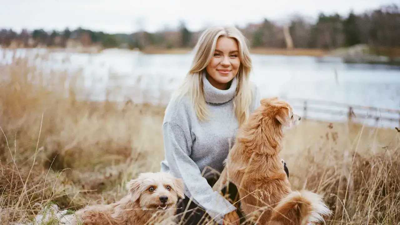 En kvinna med blont hår och en grå polotröja sitter i torrt gräs med två hundar. Detta är en del av beas liv vid vattnet.