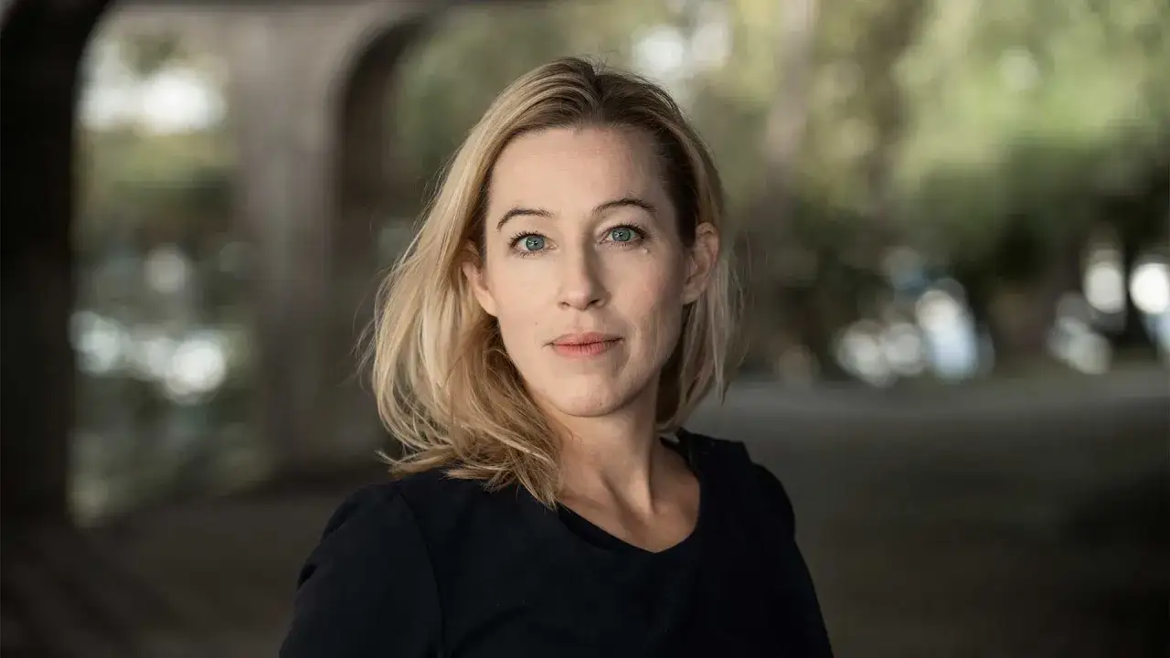 Anna Careborg, en kvinna med blont hår och blå ögon, tittar rakt in i kameran. Bakgrunden är suddig med grönska och en valvformad struktur.
