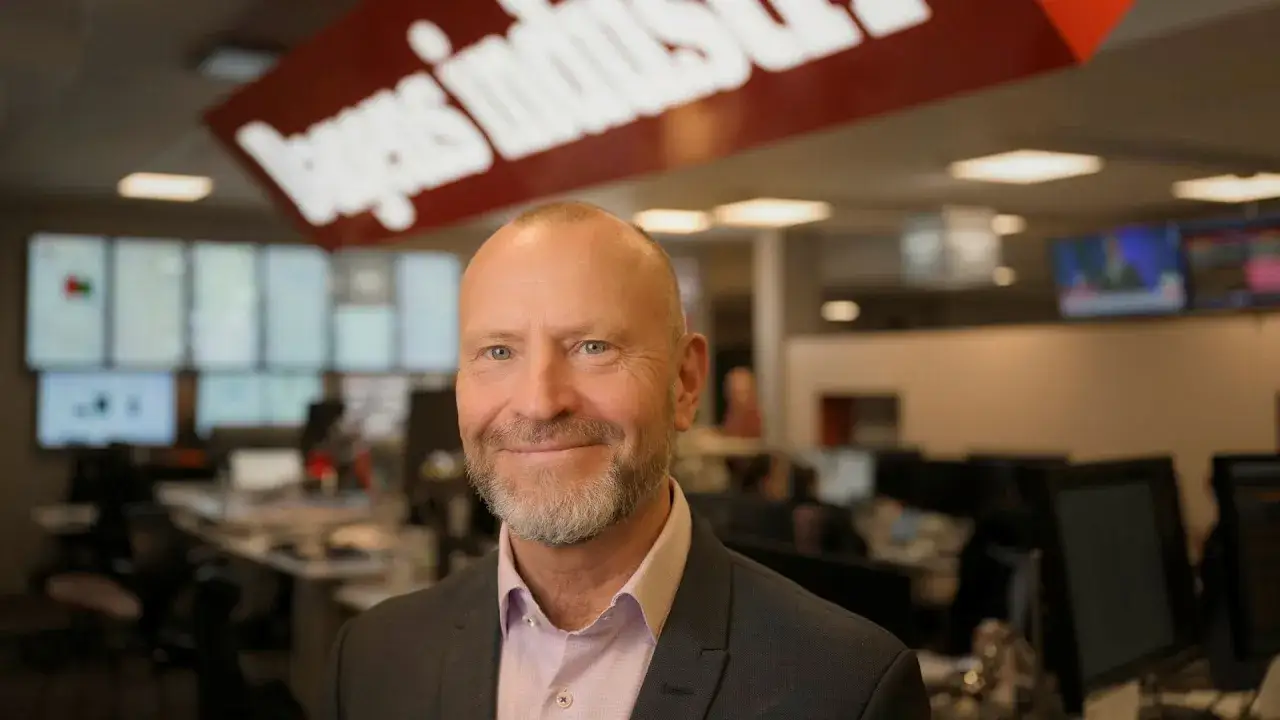 Peter Fellman ler mot kameran i en kontorsmiljö med skärmar och en röd logotyp i bakgrunden.