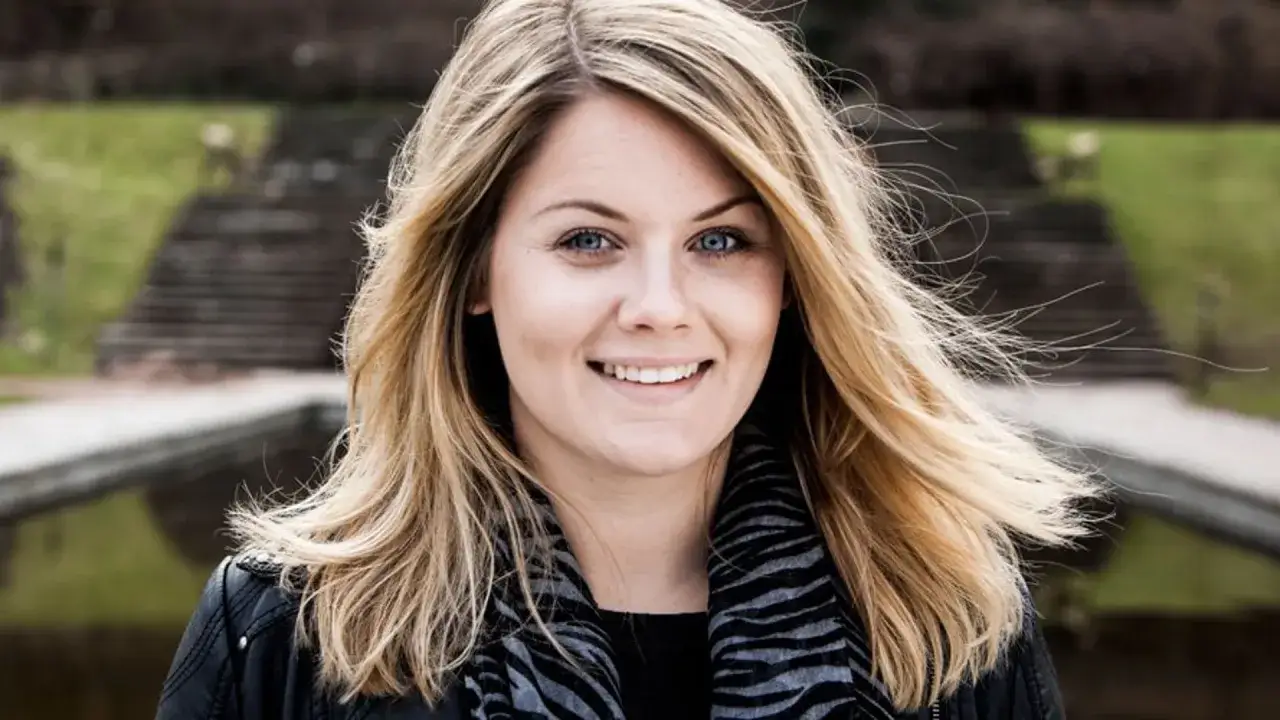Julia Karlsten ler mot kameran med blont hår som blåser i vinden. Hon bär en svart jacka och en randig halsduk.