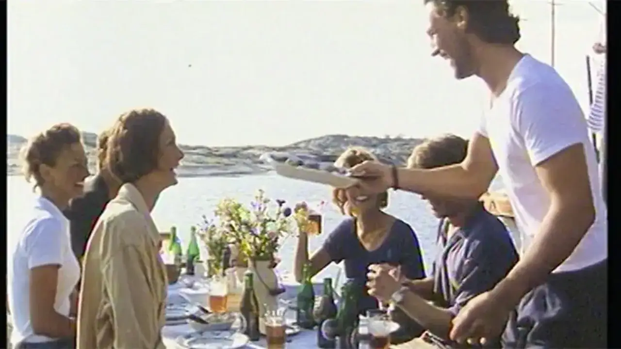 En solig sommardag vid vattnet, en festlig dukning med blommor och flaskor. En man sträcker fram en bricka, kanske med en Pripps Blå, till glada sällskap.