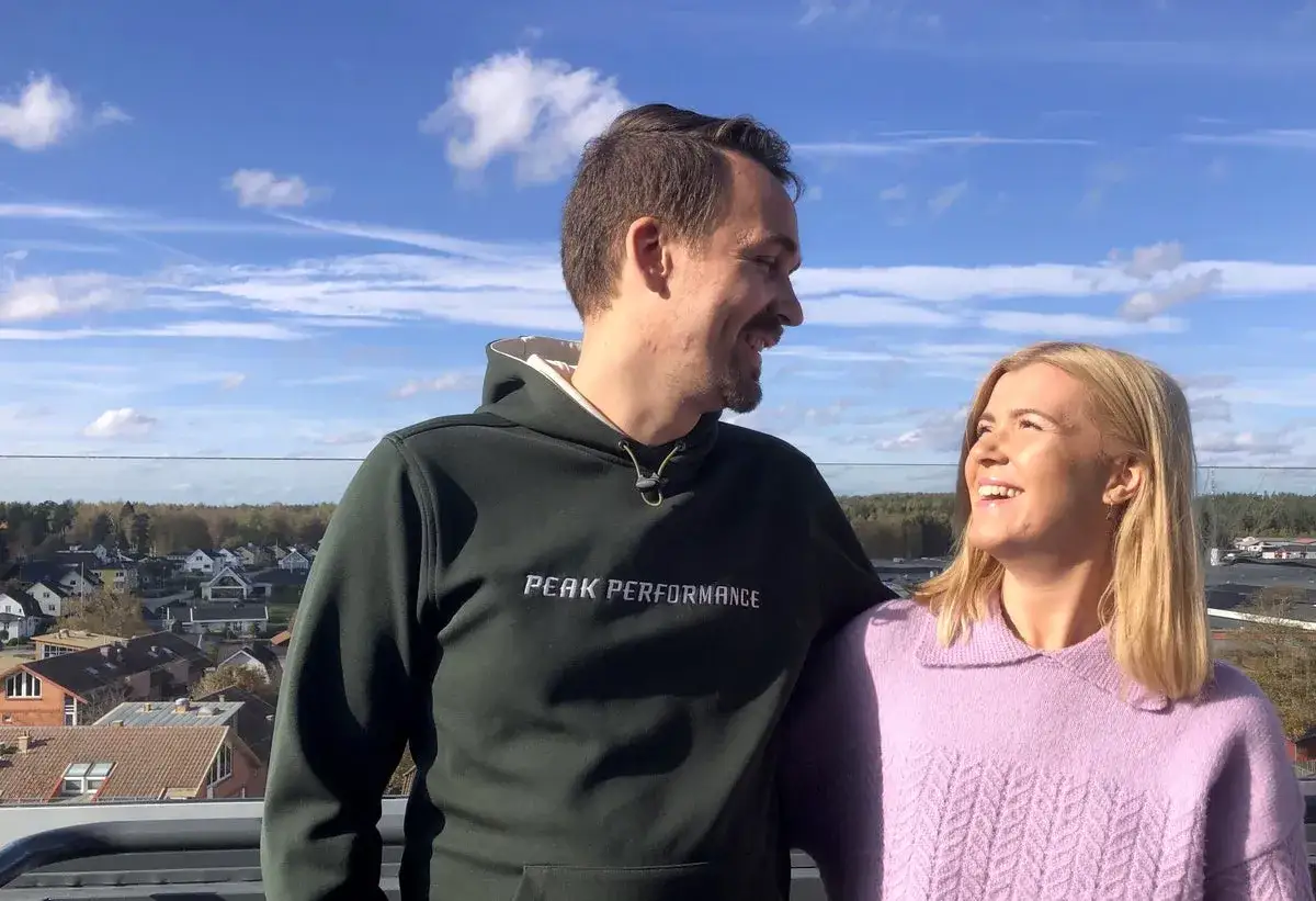 Hanna Theorin och en man ler mot varandra med en stad i bakgrunden under en blå himmel med moln.