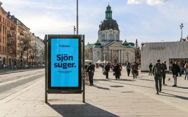 Folksam reklam på en blå skylt i en stadsmiljö. "Sjön suger" står det, med texten "Sparande och försäkring ska inte göra det." under.