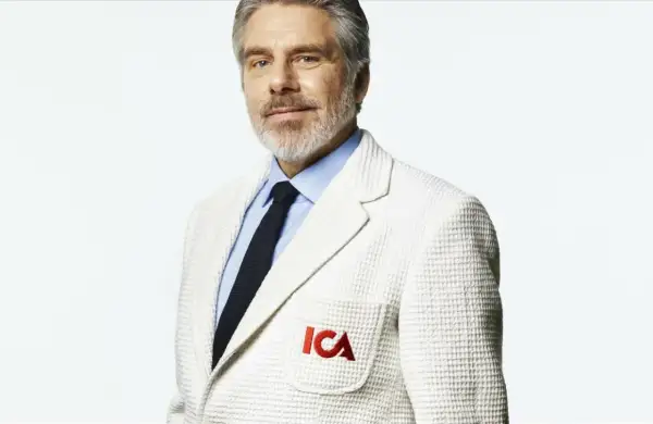 Man med grått skägg och blå skjorta, bär en vit kavaj med ICA-logga. Han ser ut att vara en del av nya ICA Stig.