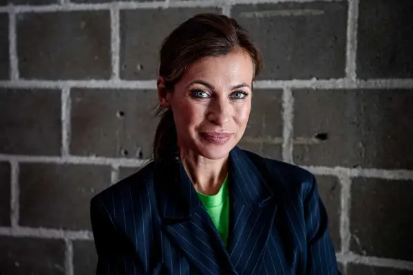 Amanda Schulman ung, med blå ögon och en grön tröja under en randig kavaj, ler mot kameran framför en tegelvägg.