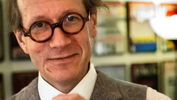 Johan Hakelius, en man med runda glasögon, ler mot kameran. Bakom honom syns bokhyllor.