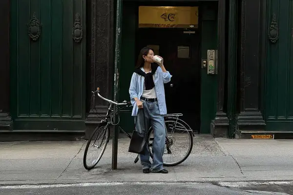 En kvinna i avslappnad street style dricker kaffe bredvid sin cykel framför en mörkgrön dörr.